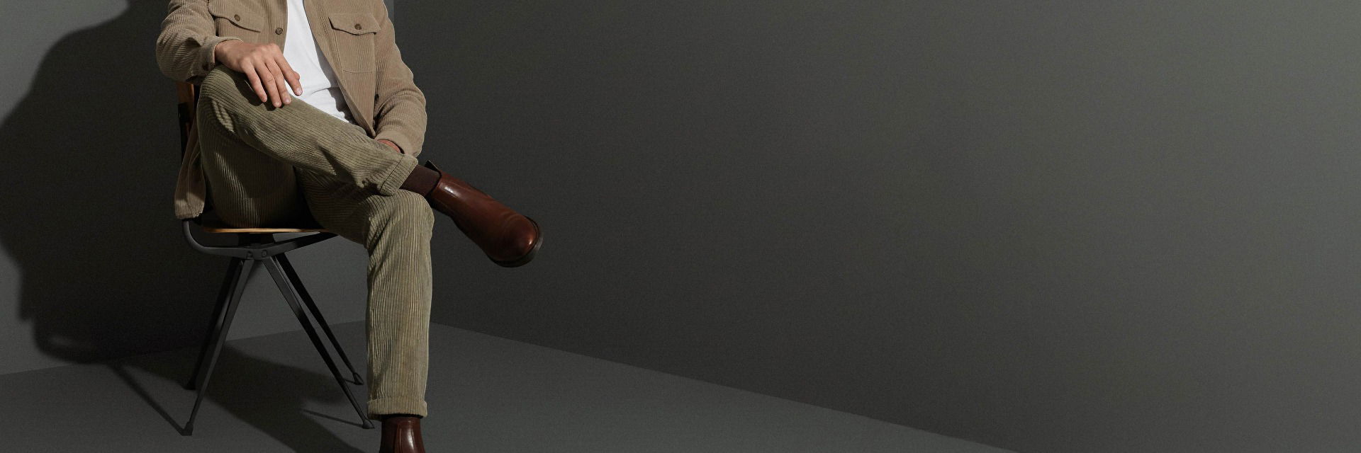 Mann sitzt auf Stuhl, trägt braune Cordhose, beige Cordjacke, weiße T-Shirt und braune Lederstiefel vor grauer Wand. Mann sitzt auf Stuhl, trägt braune Cordhose, beige Cordjacke, weiße T-Shirt und braune Lederstiefel vor grauer Wand.