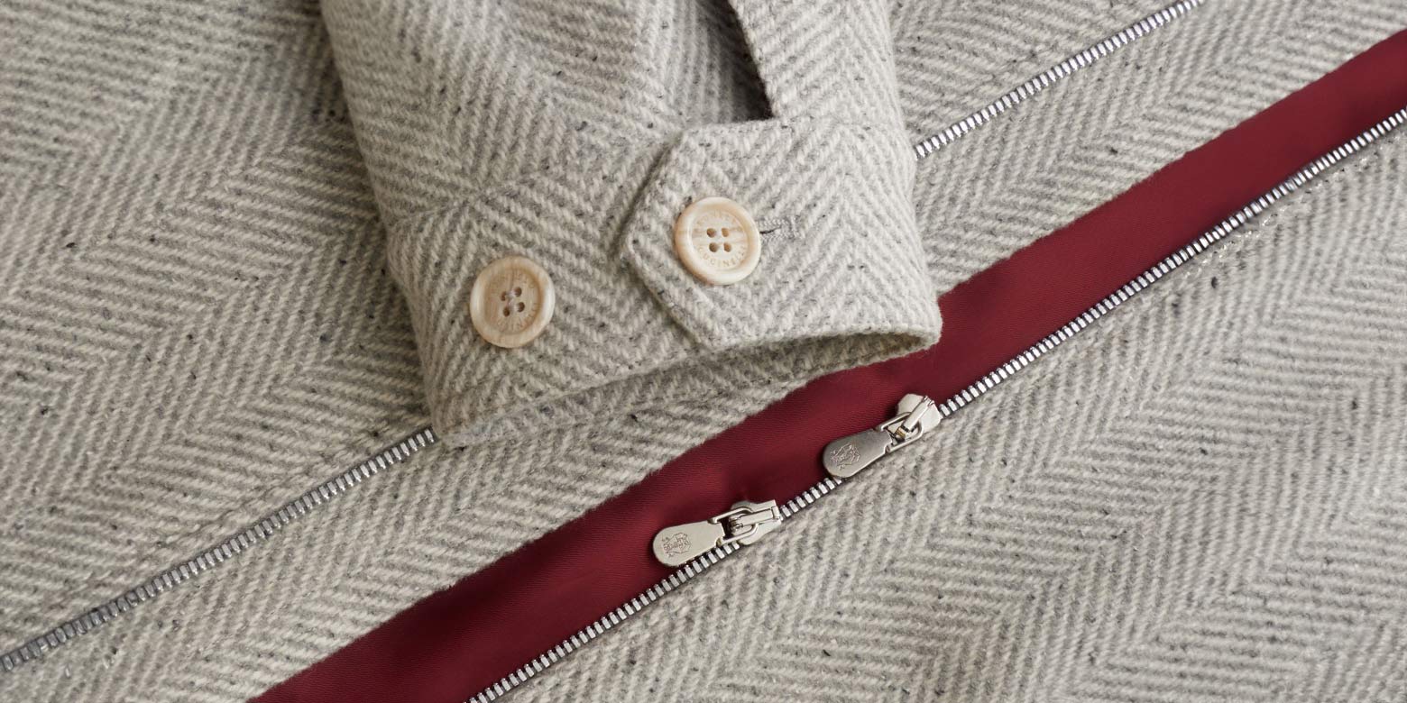 Detailaufnahme eines grau gemusterten Blousons mit silbernem Reißverschluss, rotem Innenfutter und beige Knöpfen am Ärmel.