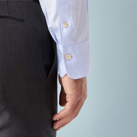 Detailaufnahme einer hellblauen Herrenhemd-Manschette mit Knöpfen am Handgelenk neben einer dunkelgrauen Hose vor hellblauem Hintergrund.