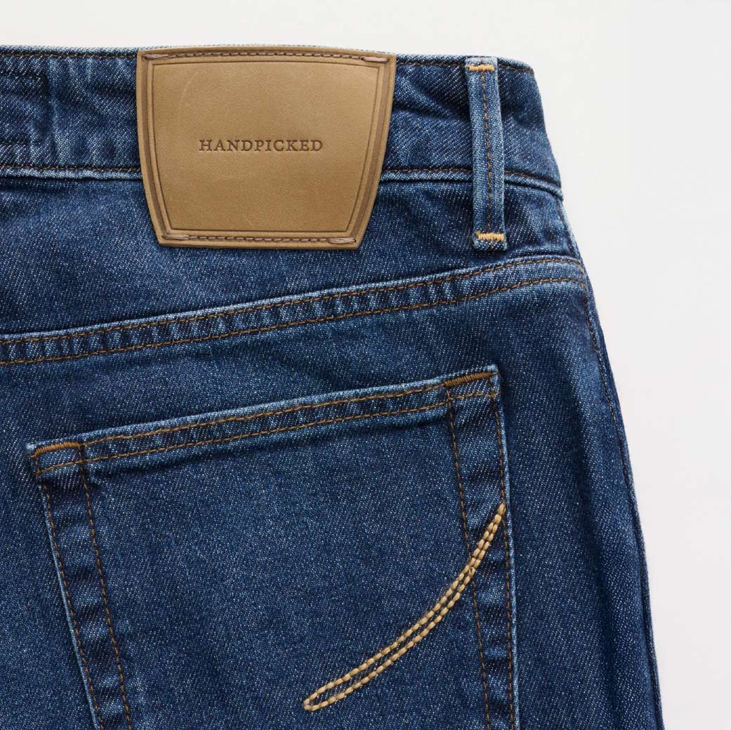 Detailansicht der Rückseite einer dunkelblauen Jeans mit braunem Lederpatch und gelber Ziernaht am Saum und Taschenrand.