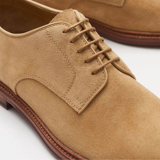 Beige Veloursleder-Schnürschuh mit feiner Naht, brauner Lederlaufsohle und klassischen Rundschnürsenkeln, Nahaufnahme. Beige Veloursleder-Schnürschuh mit feiner Naht, brauner Lederlaufsohle und klassischen Rundschnürsenkeln, Nahaufnahme.