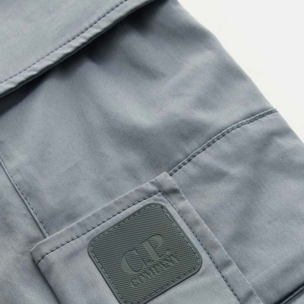 Detailaufnahme einer silbergrauen Cargohosen-Tasche mit eingenähtem CP Company Logopatch aus Gummi.