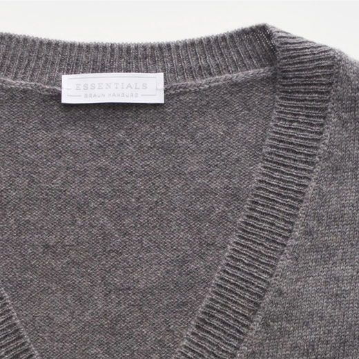 Detailaufnahme eines grauen Herrenpullunders mit V-Ausschnitt und weißem Designerlabel „ESSENTIALS BRAUN HAMBURG“ am Kragen. Detailaufnahme eines grauen Herrenpullunders mit V-Ausschnitt und weißem Designerlabel „ESSENTIALS BRAUN HAMBURG“ am Kragen.