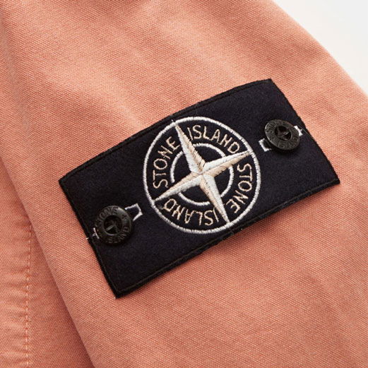 Detailaufnahme eines schwarzen Stone Island Logopatches mit Kompass-Stickerei auf hellorangefarbener Herrenjacke. Detailaufnahme eines schwarzen Stone Island Logopatches mit Kompass-Stickerei auf hellorangefarbener Herrenjacke.