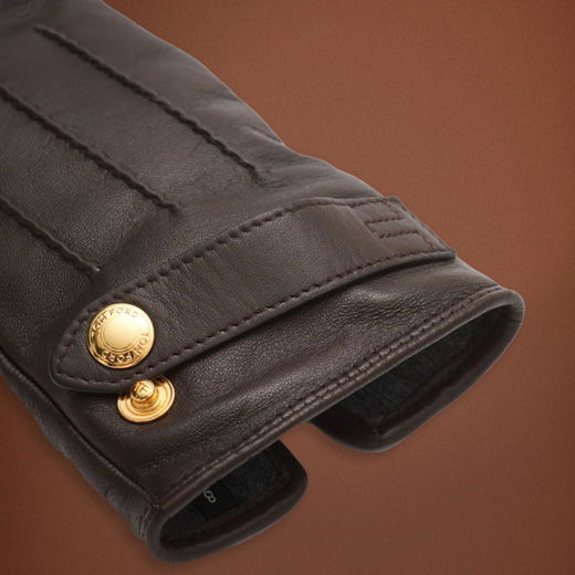 Brauner Lederhandschuh mit feiner Naht und goldfarbenem Knopf mit Gravur, Detailansicht des Handgelenks.