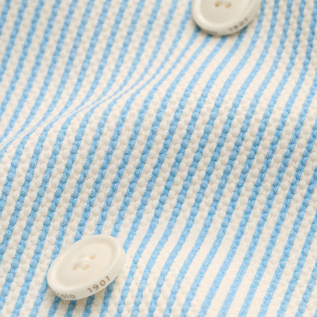 Detailansicht eines weiß-hellblau gestreiften Baumwollsakkostoffs mit zwei weißen Knöpfen.