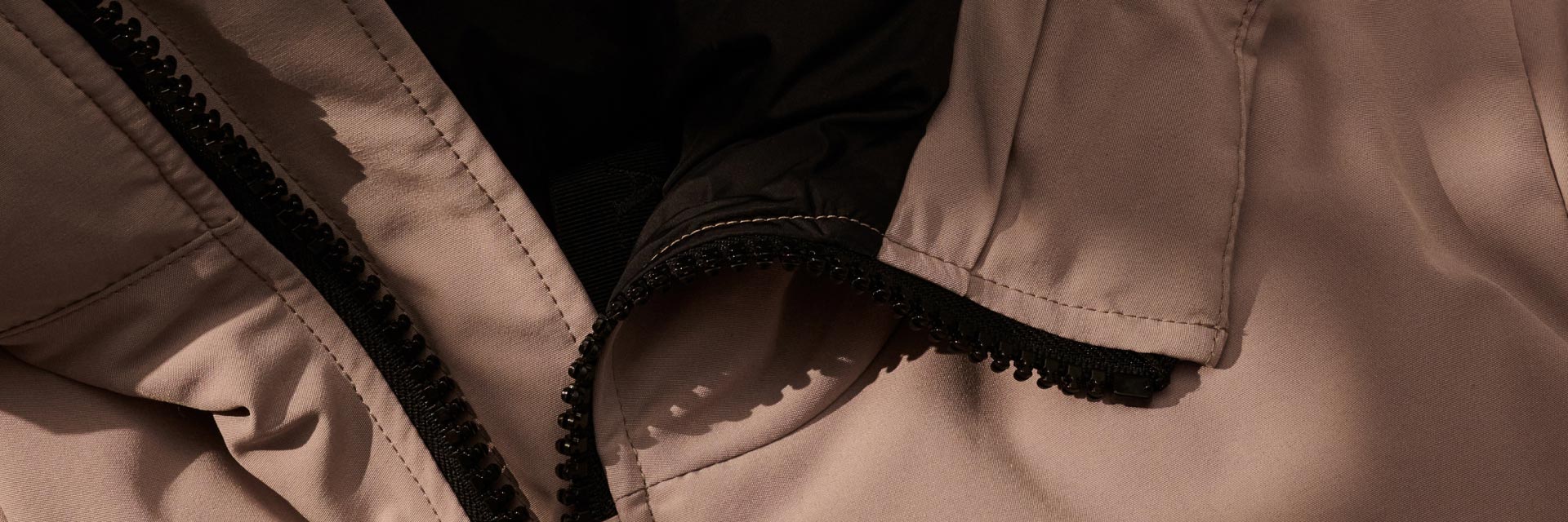 Detailaufnahme des schwarzen Reißverschlusses einer hellbraunen Herren-Regenjacke mit sichtbaren Nähten und Stoffstruktur. Detailaufnahme des schwarzen Reißverschlusses einer hellbraunen Herren-Regenjacke mit sichtbaren Nähten und Stoffstruktur.