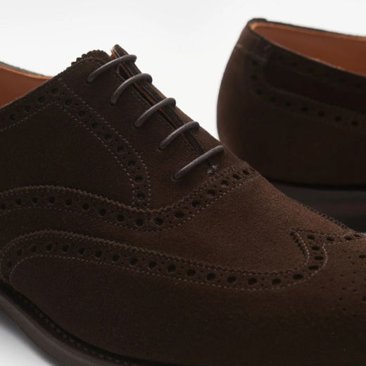 Dunkelbraune Herren-Brogues aus Veloursleder mit dekorativen Lochmustern und braunen Schnürsenkeln, Nahaufnahme.
