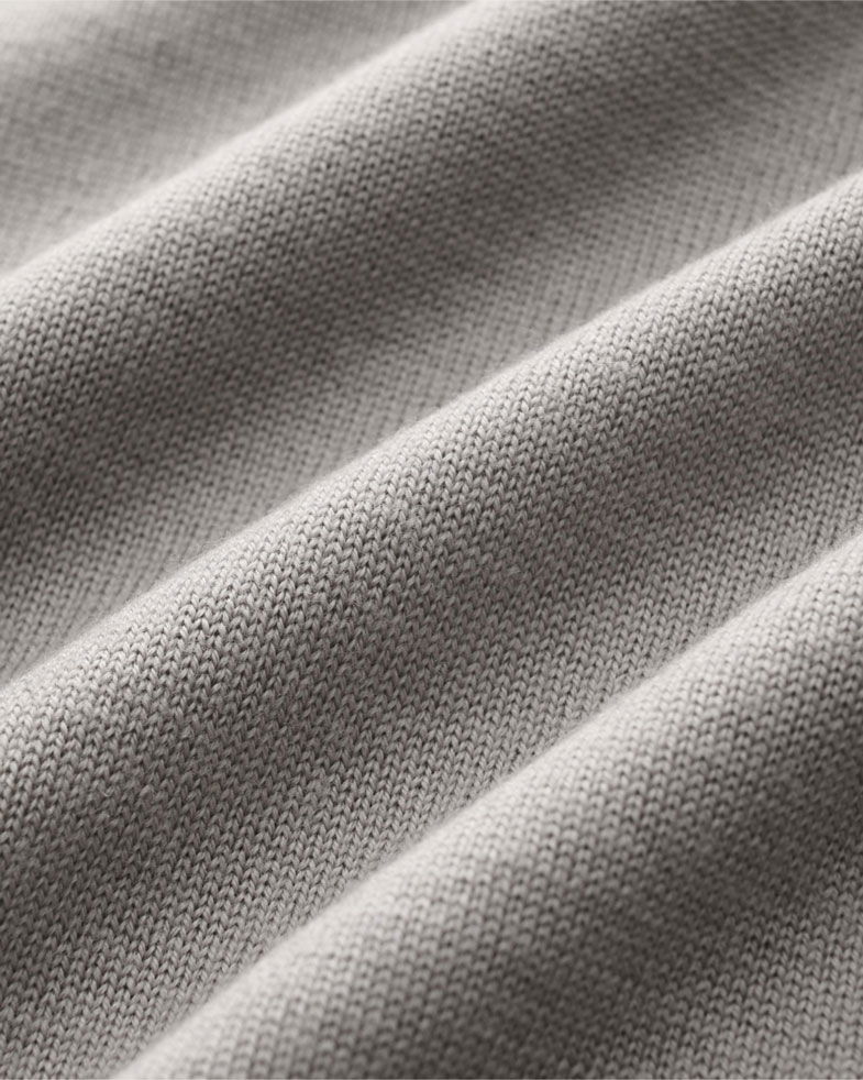 Nahaufnahme eines weich strukturierten Wollstoffes in Grau-Beige mit feiner, gleichmäßiger Maschenstruktur und Faltenwurf.