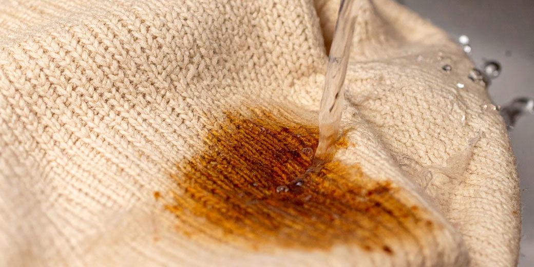 Beiger Pullover mit Nahaufnahme eines Rostflecks, der von einem Wasserstrahl überspült wird, sichtbare Textur des Strickstoffs.