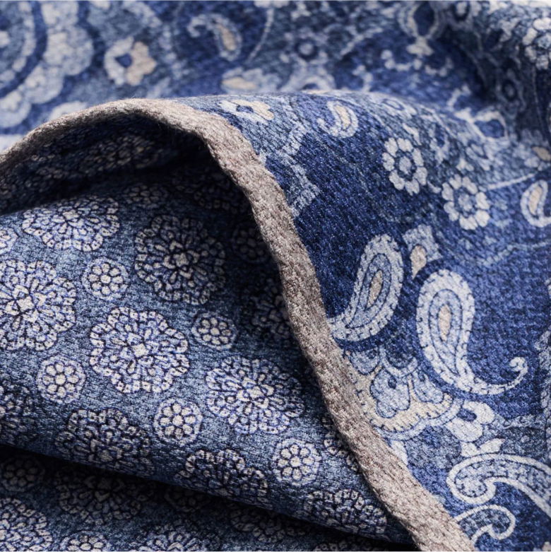 Detailaufnahme eines blau-weiß-beige gemusterten Stoffes mit floralen und Paisley-Motiven und grauem Saum.