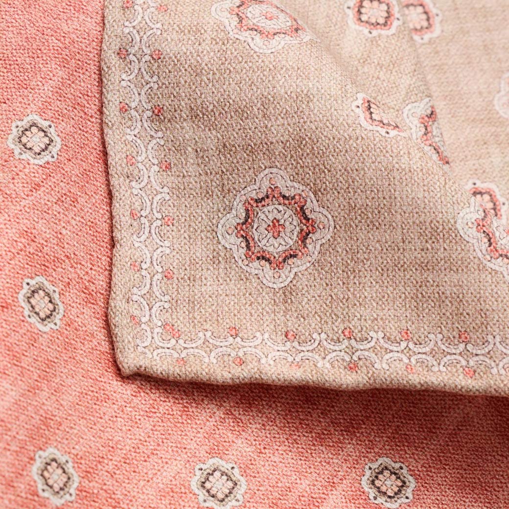 Beige-orange Einstecktuch mit floralen Mustern und dekorativem Rand, auf orange gemustertem Stoff liegend.
