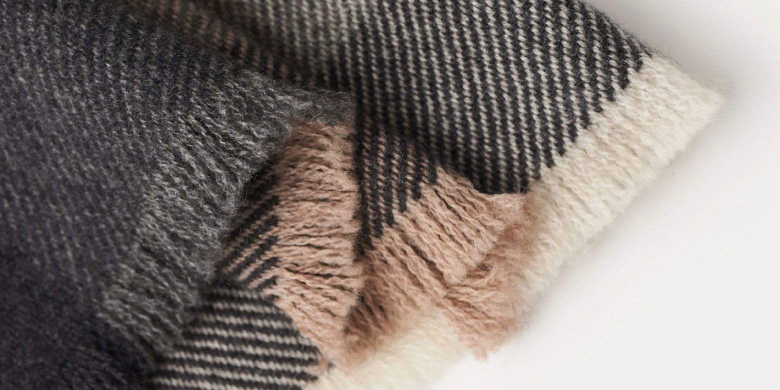 Detailaufnahme eines karierten Schals in Dunkelgrau, Braun und Creme mit weichem, flauschigem Fransensaum auf weißem Hintergrund.