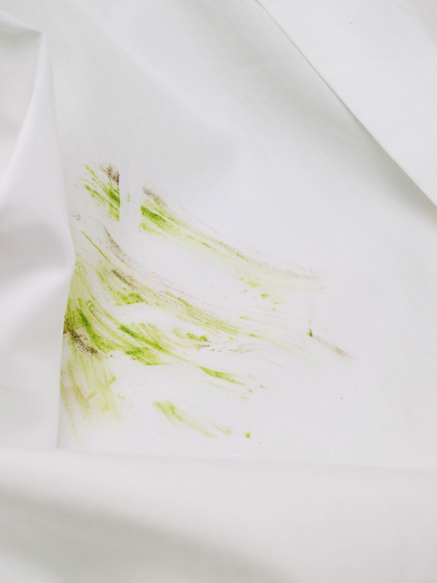 Weißer Hemdstoff mit deutlichen grünen Grasflecken und leichten braunen Verschmutzungen, Detailaufnahme.