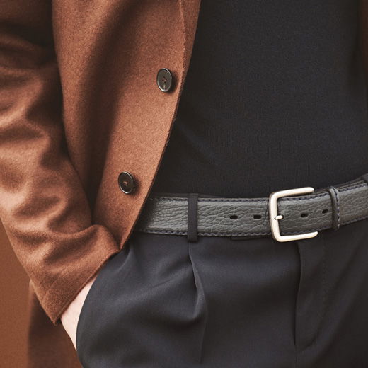 Brauner Mantel, schwarzer Pullover, schwarze Hose mit schwarzem Ledergürtel und silberner Schnalle, Hand in Tasche.