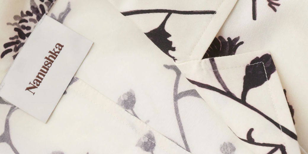 Detailaufnahme eines weiß-braun gemusterten Kurzarmhemdes mit sichtbarem Nanushka-Designerlabel am Kragen. Detailaufnahme eines weiß-braun gemusterten Kurzarmhemdes mit sichtbarem Nanushka-Designerlabel am Kragen.