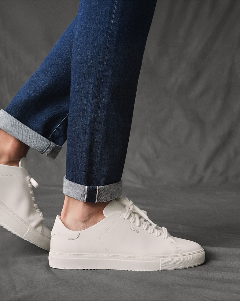 Detailaufnahme einer dunkelblauen Selvedge-Denim-Jeans mit Krempelung, getragen mit weißen Arigato-Sneakern.