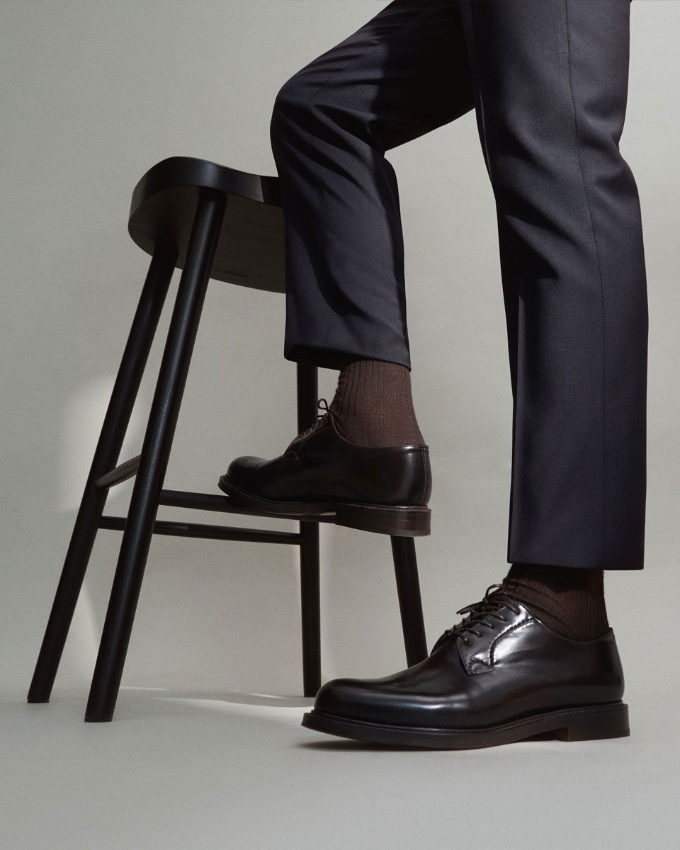 Detailaufnahme von braunen Socken mit schwarzen Schnürschuhen und dunkelblauer Hose, ein Fuß auf einem schwarzen Stuhl. Detailaufnahme von braunen Socken mit schwarzen Schnürschuhen und dunkelblauer Hose, ein Fuß auf einem schwarzen Stuhl.