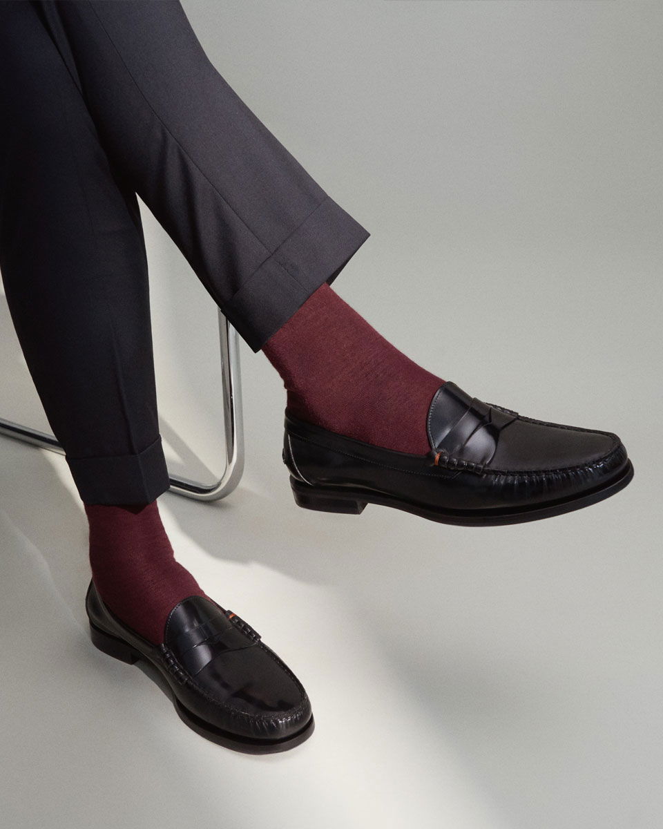 Herrenbeine in dunkelgrauer Hose, rote Socken und schwarze Loafer, sitzend auf einem Stuhl vor hellgrauem Hintergrund. Herrenbeine in dunkelgrauer Hose, rote Socken und schwarze Loafer, sitzend auf einem Stuhl vor hellgrauem Hintergrund.