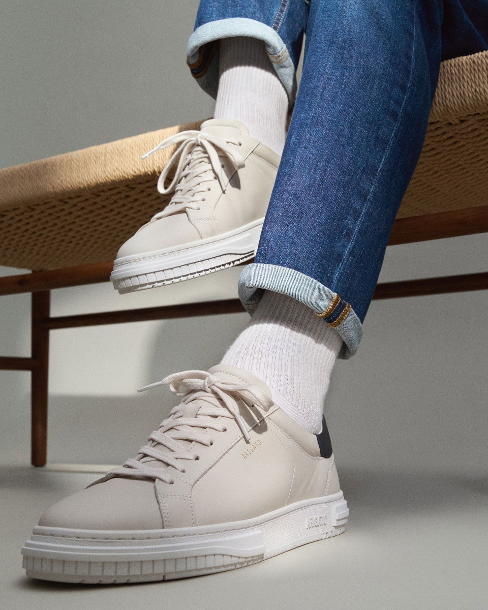 Weiß-beige Sneaker mit weißen Socken, blaue Jeans, sitzende Person auf geflochtenem Holzstuhl, Nahaufnahme. Weiß-beige Sneaker mit weißen Socken, blaue Jeans, sitzende Person auf geflochtenem Holzstuhl, Nahaufnahme.