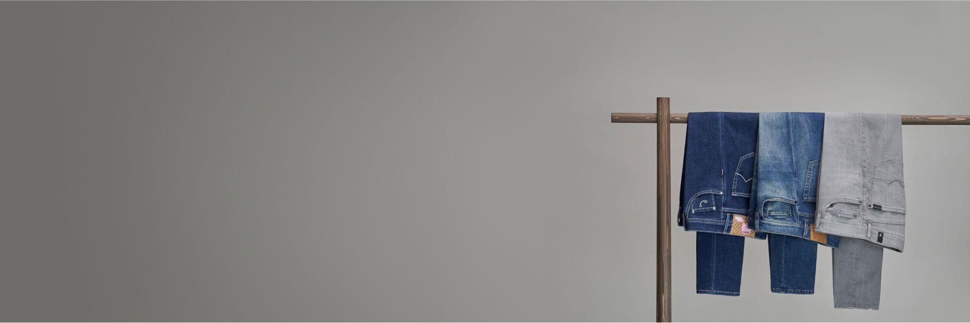 Drei Herrenjeans in Dunkelblau, Hellblau und Grau hängen ordentlich auf einer Holzstange vor grauem Hintergrund. Drei Herrenjeans in Dunkelblau, Hellblau und Grau hängen ordentlich auf einer Holzstange vor grauem Hintergrund.