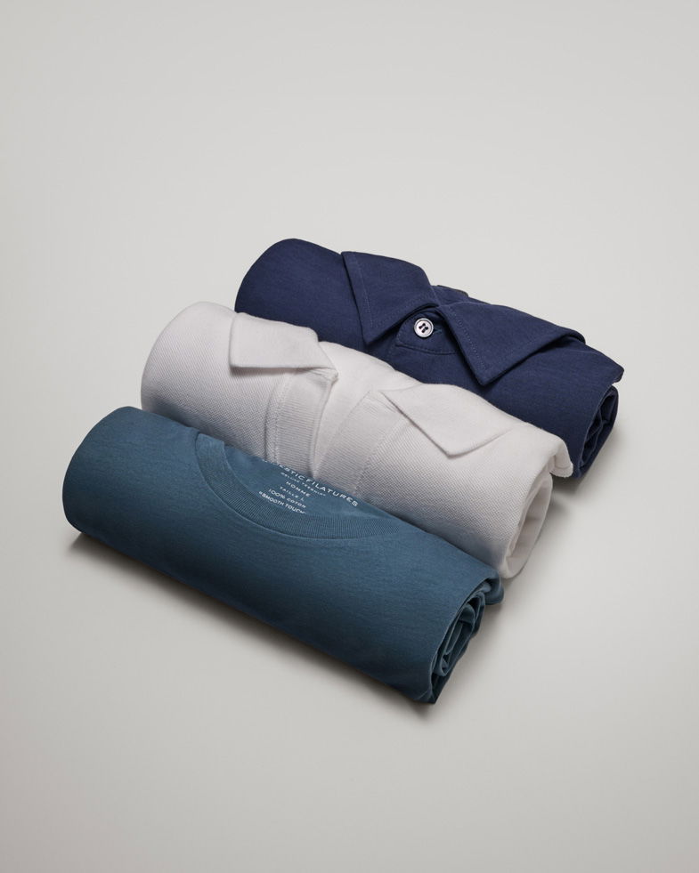 Drei eingerollte Herren-Shirts in Dunkelblau, Hellgrau und Grün liegend auf hellem Untergrund.