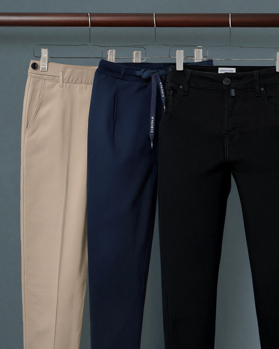 Drei hängende Herrenhosen in Beige, Navy mit Kordelzug und Schwarz, an silbernen Kleiderbügeln vor grauem Hintergrund.