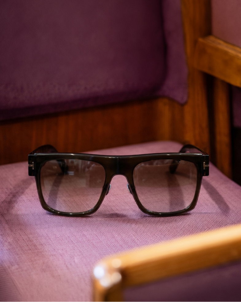 Schwarze eckige Acetat-Sonnenbrille mit abgetönten Gläsern liegt auf violettem Stuhlpolster aus Holz.