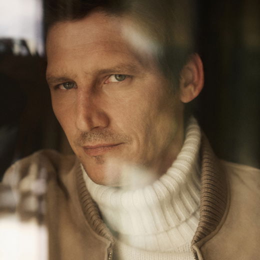 Ein Mann mit kurzem braunem Haar trägt einen weißen Rollkragenpullover und eine beige Lederjacke, schaut direkt in die Kamera. Ein Mann mit kurzem braunem Haar trägt einen weißen Rollkragenpullover und eine beige Lederjacke, schaut direkt in die Kamera.