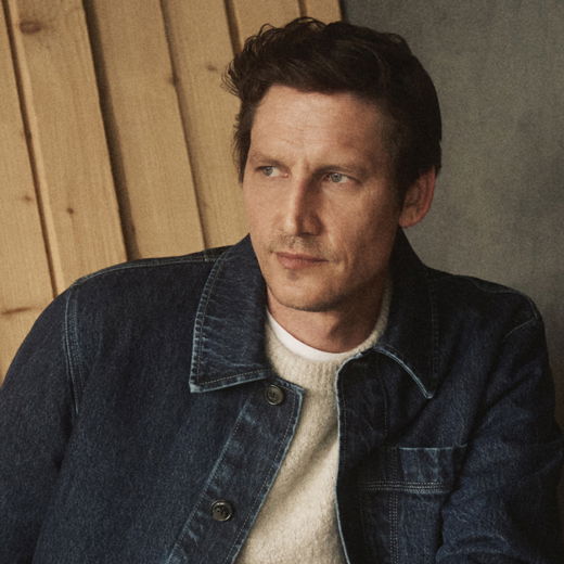 Herrenportrait: Mann mit beigem Strickpullover und blauer Jeansjacke schaut nachdenklich seitlich, Holz- und Betonwand im Hintergrund. Herrenportrait: Mann mit beigem Strickpullover und blauer Jeansjacke schaut nachdenklich seitlich, Holz- und Betonwand im Hintergrund.