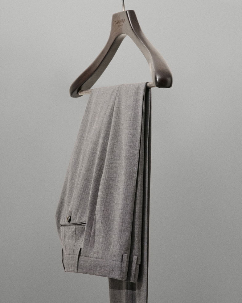 Grau melierte Herrenhose mit Bügelumschlag, ordentlich auf einem braunen Holzbügel vor schlicht grauem Hintergrund hängend.