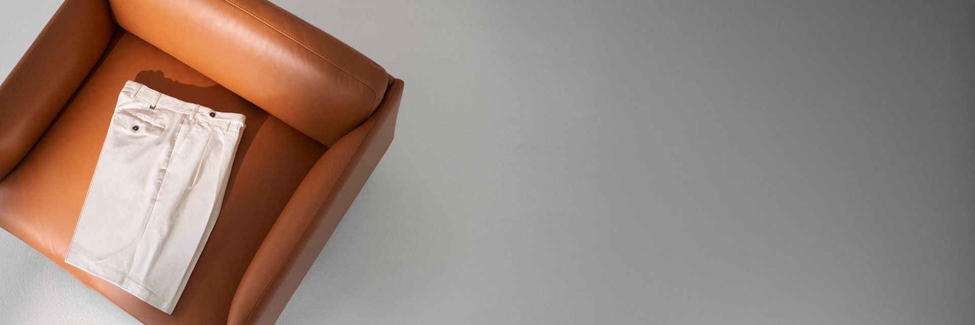 Beige Herren-Shorts ordentlich gefaltet auf einem braunen Ledersessel, von oben fotografiert, grauer Hintergrund. Beige Herren-Shorts ordentlich gefaltet auf einem braunen Ledersessel, von oben fotografiert, grauer Hintergrund.