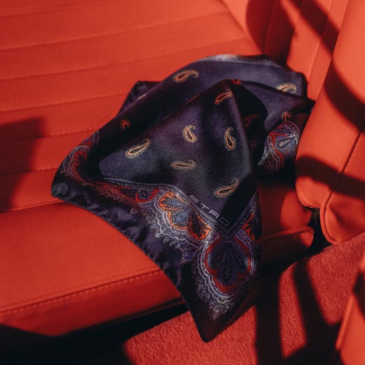 Ein navyblaues Einstecktuch mit Paisley-Muster liegt auf einem roten Ledersitz im Auto, Sonnenlicht fällt darauf. Ein navyblaues Einstecktuch mit Paisley-Muster liegt auf einem roten Ledersitz im Auto, Sonnenlicht fällt darauf.