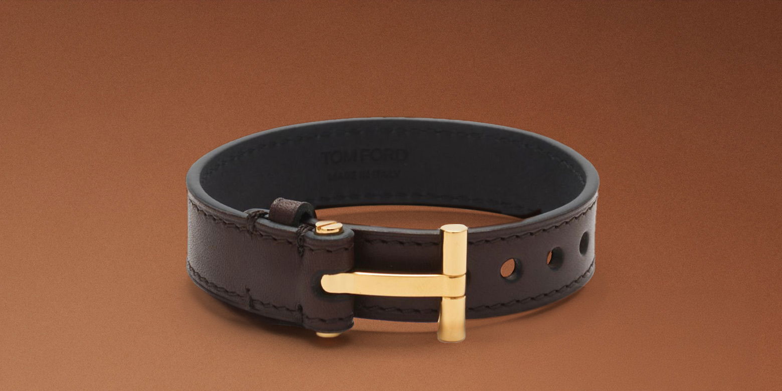 Braunes Herren-Lederarmband mit goldener Schnalle in Form eines Designerlogos, freistehend vor braunem Hintergrund.