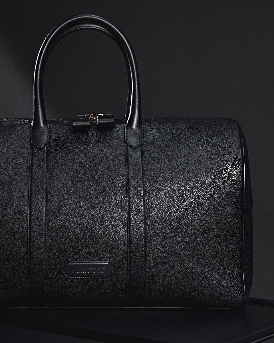 Schwarze Reisetasche aus genarbtem Leder mit zwei Henkeln und sichtbarem Tom Ford Designerlogo vor schwarzem Hintergrund.