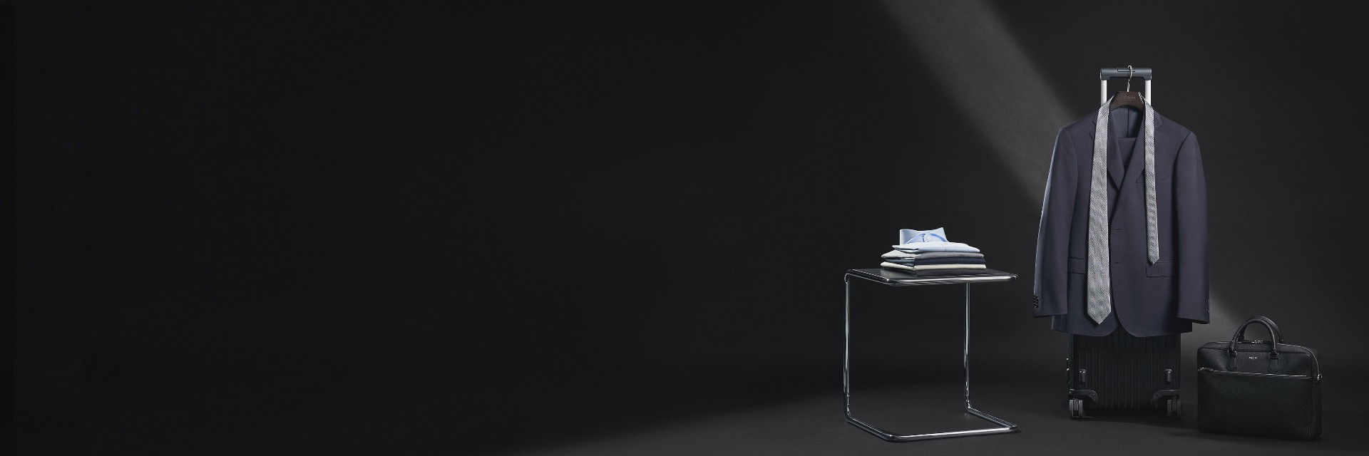 Eleganter navy Anzug mit gemusterter Krawatte an Koffer, daneben gefaltete Hemden auf modernem Hocker und schwarze Aktentasche. Eleganter navy Anzug mit gemusterter Krawatte an Koffer, daneben gefaltete Hemden auf modernem Hocker und schwarze Aktentasche.