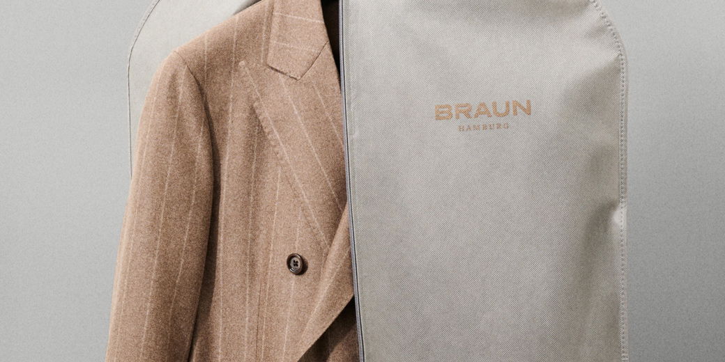 Braun-weiß gestreiftes Sakko, halb in grauem Kleidersack mit Aufschrift 'BRAUN HAMBURG' hängend vor grauem Hintergrund.