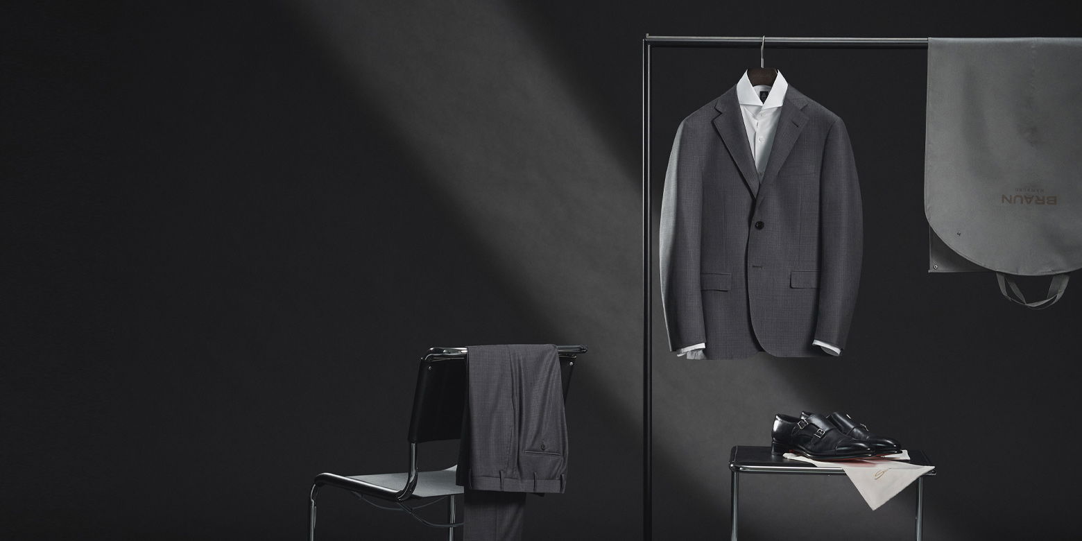 Graues Sakko mit weißem Hemd am Kleiderbügel, graue Hose über Stuhl, schwarze Monks auf Hocker, Kleidersack rechts.