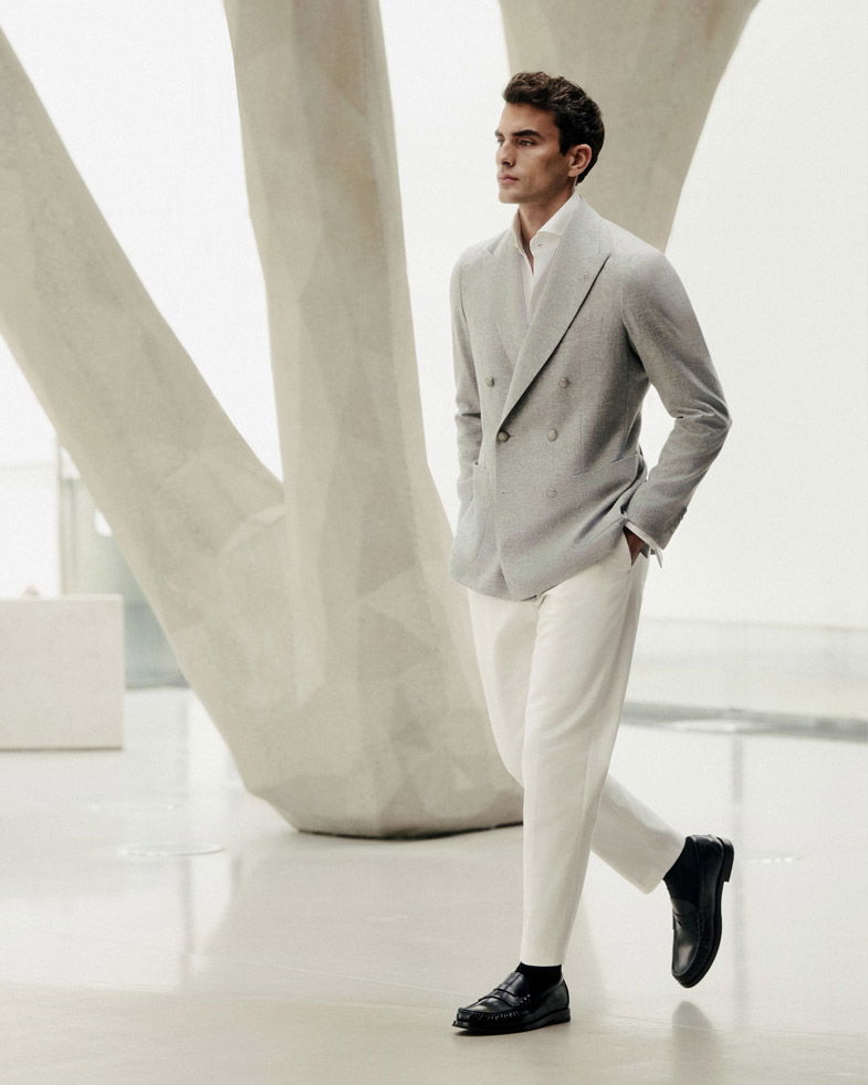 Herr geht in hellgrauem Sakko, weißem Hemd, weißer Hose und schwarzen Loafern vor hellem, modernem Hintergrund. Herr geht in hellgrauem Sakko, weißem Hemd, weißer Hose und schwarzen Loafern vor hellem, modernem Hintergrund.