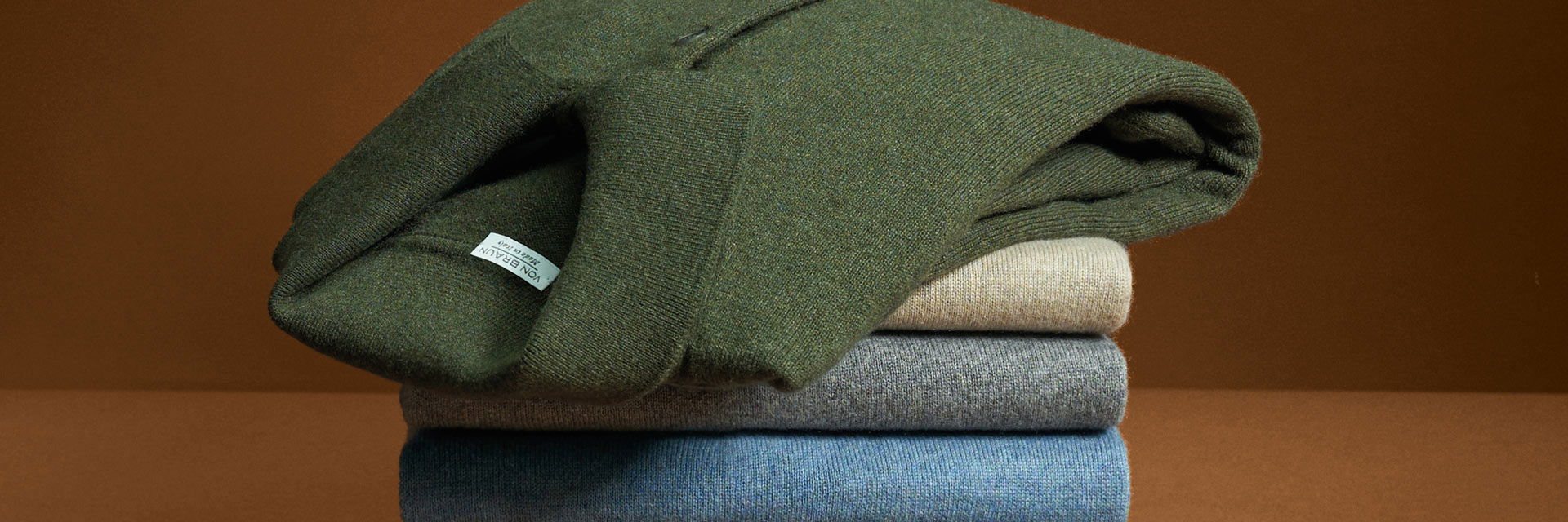 Gestapelte Herren-Wollpullover in verschiedenen Farben, oben ein grüner Pullover mit sichtbarem Etikett vor braunem Hintergrund. Gestapelte Herren-Wollpullover in verschiedenen Farben, oben ein grüner Pullover mit sichtbarem Etikett vor braunem Hintergrund.