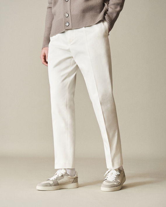 Herren trägt weiße Hose, weiße Sneaker mit grauen Details und hellbraunen Cardigan mit Knöpfen, Hände in Taschen.