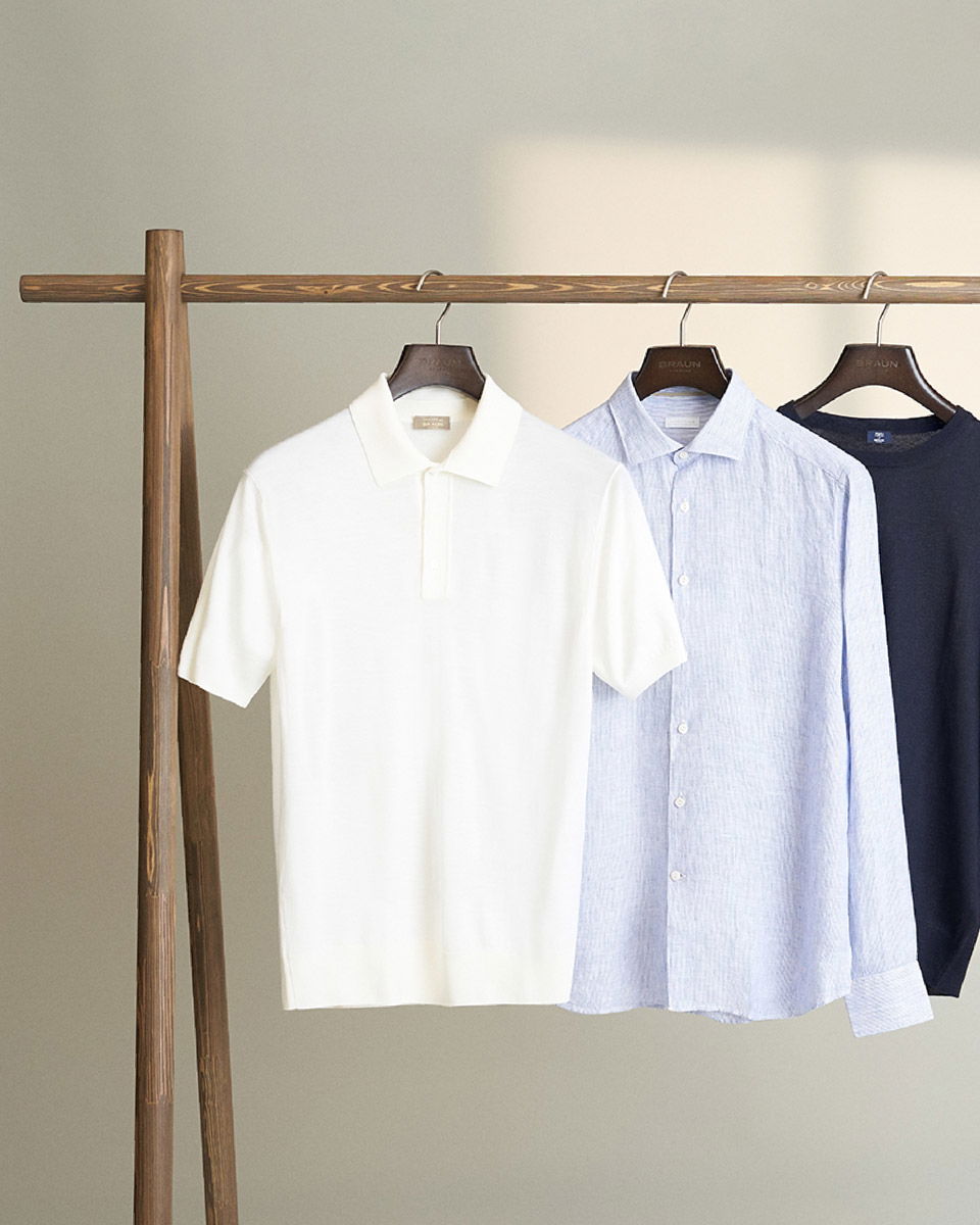 Weiße Polo, hellblaues Hemd und dunkelblauer Pullover hängend an Holzkleiderstange vor beigem Hintergrund.