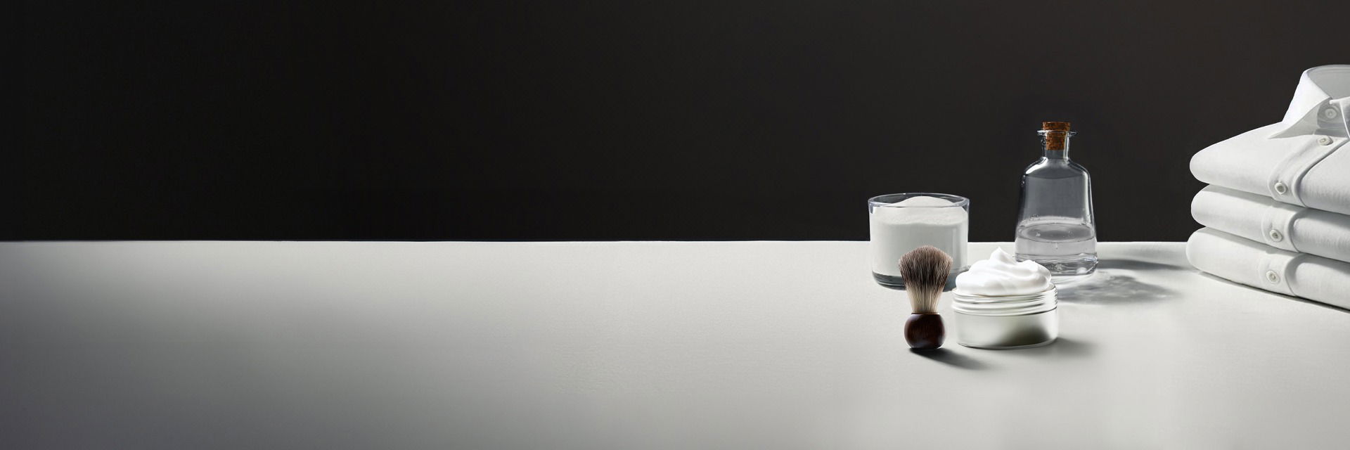 Weißer Tisch mit gestapelten weißen Hemden, Glas mit Backpulver, Rasierschaumpinsel, Rasierschaum und Flasche Essig vor schwarzem Hintergrund. Weißer Tisch mit gestapelten weißen Hemden, Glas mit Backpulver, Rasierschaumpinsel, Rasierschaum und Flasche Essig vor schwarzem Hintergrund.