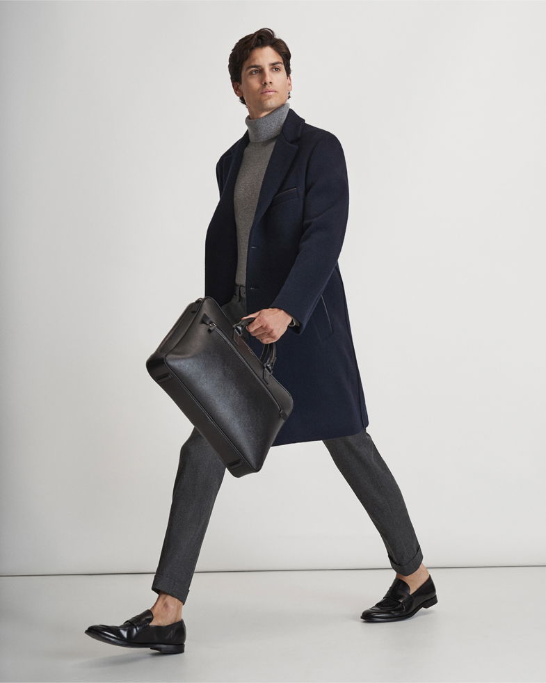 Mann trägt dunkelblauen Mantel, grauen Rollkragenpullover, graue Hose, schwarze Loafer und hält schwarze Aktentasche. Mann trägt dunkelblauen Mantel, grauen Rollkragenpullover, graue Hose, schwarze Loafer und hält schwarze Aktentasche.
