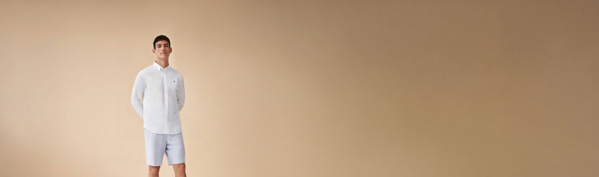 Junger Mann steht vor sandfarbenem Hintergrund, trägt weißes Hemd und hellblaue Shorts, Hände hinter dem Rücken. Junger Mann steht vor sandfarbenem Hintergrund, trägt weißes Hemd und hellblaue Shorts, Hände hinter dem Rücken.
