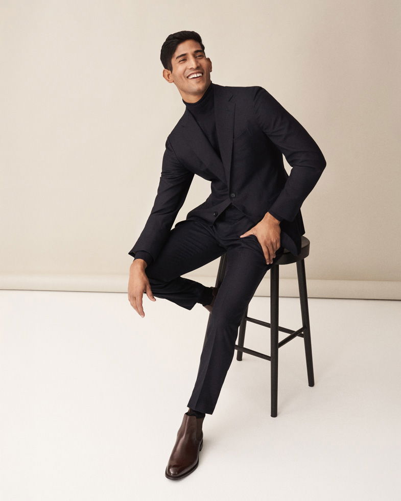 Mann sitzt auf schwarzem Hocker, trägt schwarzen Anzug, schwarzen Rollkragenpullover und braune Chelsea-Boots aus Glattleder.