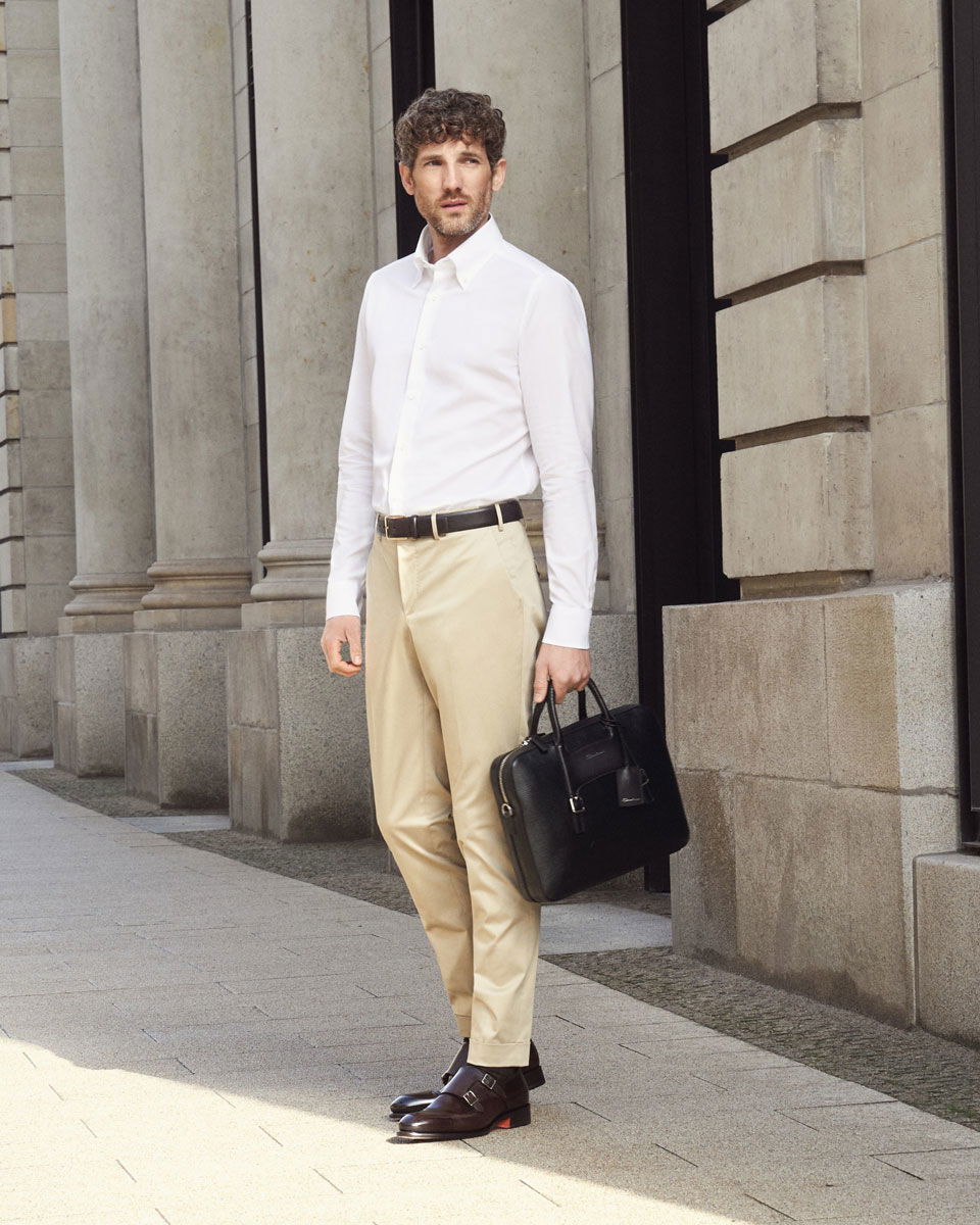 Mann mit weißem Businesshemd, beiger Chinohose, braunen Doppelmonks und schwarzer Aktentasche vor Säulengebäude stehend.