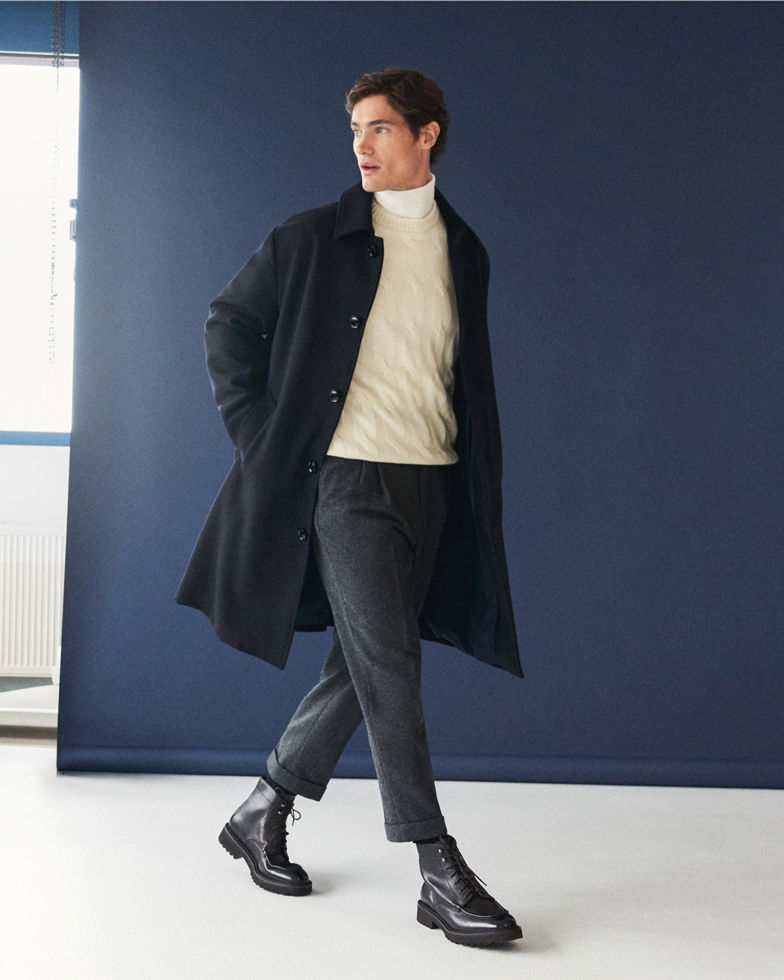 Mann trägt dunkelblauen Mantel, cremefarbenen Rollkragenpullover, dunkelgraue Cordhose und schwarze Schnürboots, geht nach links.