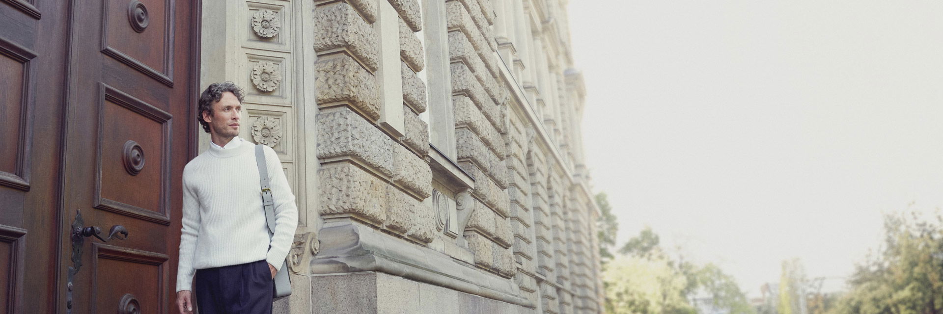 Mann trägt weißes Hemd, cremefarbigen Rundhalspullover, dunkelblaue Hose und hellgraue Tasche vor historischem Gebäude. Mann trägt weißes Hemd, cremefarbigen Rundhalspullover, dunkelblaue Hose und hellgraue Tasche vor historischem Gebäude.