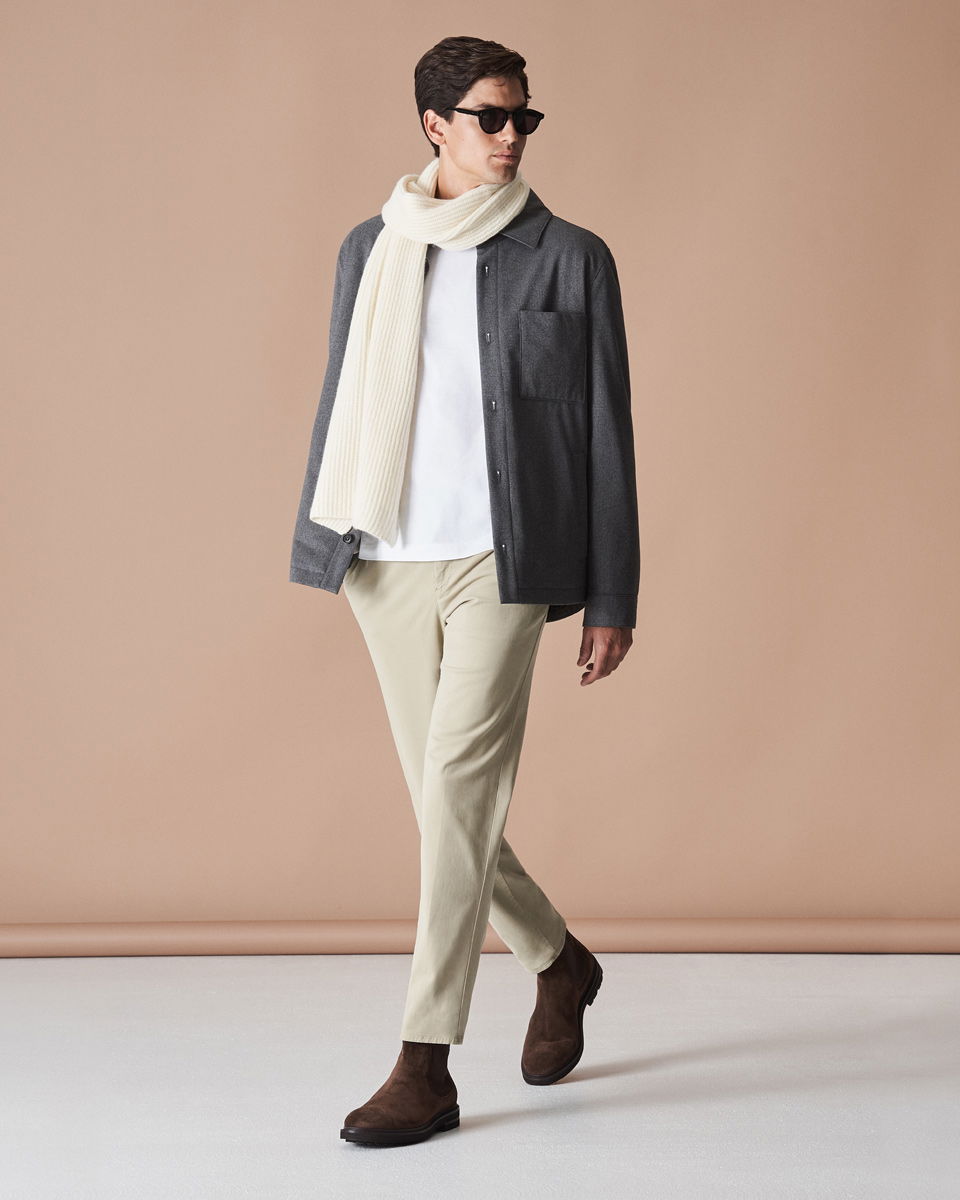 Mann mit weißem T-Shirt, grauem Overshirt, beiger Hose, cremefarbenem Schal, braunen Boots und schwarzer Sonnenbrille vor beigem Hintergrund. Mann mit weißem T-Shirt, grauem Overshirt, beiger Hose, cremefarbenem Schal, braunen Boots und schwarzer Sonnenbrille vor beigem Hintergrund.